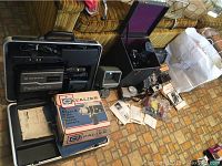 Photo showing vintage film equipment: Panasonic VHS camcorder in case, Cavalier Super 8 camera boxes, Moviscop viewer/editor, Keystone 16mm projector, and related accessories on floor