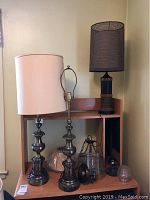 Three vintage table lamps on a wooden shelving unit, one with dark mesh shade, two with cream fabric shades; also metal and glass ceiling fixture and wall sconce visible below lamps.