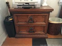 Full front view showing the bedside table on a hardwood floor with a printer on top and wastebasket to the side.