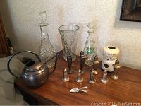 Sterling silver baby spoon, weighted sterling salt and pepper shakers in four sets, two glass decanters, glass vase, ceramic candle holder and decorative metal oil can on wooden surface.