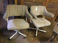 Pair of beige upholstered metal rolling chairs with armrests and caster wheels, showing wear and paint loss in a room with carpet and wooden door shutters.
