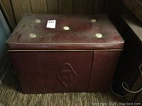 Front and top angled view of burgundy vinyl padded storage chest with tufted lid showing large round buttons and surface wear.