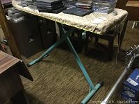 Side view of vintage turquoise metal folding ironing board with patterned padded top and rubber foot caps on legs
