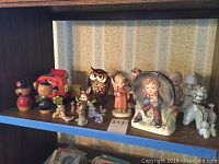 Photo of shelf with various small ceramic figurines including Story Town piggy banks, Hummel reproductions, and poodle figurines