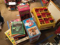 Lot of boxed board games and toys including Professor Owl Play Desk game, Dancerella doll, and Zoo books. Shows colorful items stacked on carpeted floor.