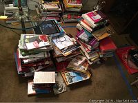 Wide view of multiple stacks of books and audiobooks on carpeted floor showing large quantity of titles and genres.
