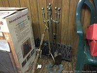 Photo of brass tone fireplace tool set with stand and fishing rod leaning against wooden wall.