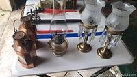 Five lighting items on a table: two copper lanterns, one oil lamp with clear glass chimney, and two brass hurricane candle holders with frosted glass lampshades and prisms.