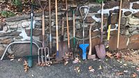 Wide view showing various garden tools lined up against a stone wall on concrete; includes bow saw, axes, spade, rakes, hoes, and plant hole digger.