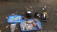 Photo of assorted hand tools including wrenches on a blue plastic lid, two black pumps, and a yellow and black trolley jack on pavement with scattered leaves.