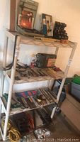 Four-tier storage shelf holding multiple hand tools, power tools, and accessories in a garage environment. Visible tools include a handsaw, files, screwdrivers, hammers, extension cords, and levels.