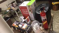 Full view of all items together on and around a table including breadmaker box, food mill, air fryer box, food processor, mandolin slicer, vegetable slicer, and fire extinguisher.