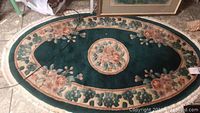 Full view of an oval floral area rug with dark green and cream colors, showing overall design and condition including fringe damage.