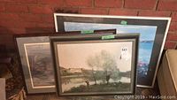 Front view of three framed outdoor scene paintings leaning against a brick wall alongside a couch.