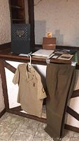 Photo showing military uniform (short-sleeve shirt with patches and medals) and gabardine wool pants hanging below a wooden counter alongside military memorabilia and documents.