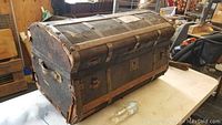 Side view of the domed antique steamer trunk showing leather reinforcements, metal latches, and handles. Trunk appears sturdy but worn.