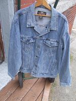 Front view of light blue denim jacket hung on a wooden hanger. Shows button front, two chest flap pockets with buttons, long sleeves, and traditional denim seams.