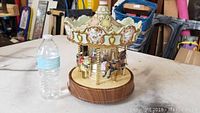 Full view of the porcelain carousel music box with scalloped canopy, multiple porcelain horses, and wood grain style base, placed next to a water bottle for scale.