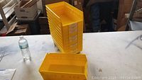 Stack of 10 yellow plastic sorting bins on a table with one additional bin lying in front. Water bottle nearby for scale.