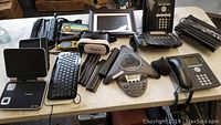Overview photo showing various electronics on a table including phones, keyboards, GPS, mouse, car TV screens, electronic picture frame, conference phone.