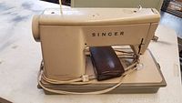 Beige Singer sewing machine with power cord, brown accessory pouch on base.