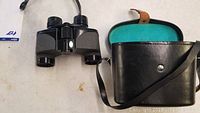 Swift binoculars with black textured grips and central focusing wheel next to their black leather carrying case with teal interior lining.