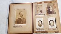 Open antique photo album with a mix of male and female portrait photographs in sepia tones, showing aged paper and captions below images.