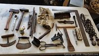 Wide view of assortment of antique hand tools and tow chain including hammers, hatchet, meat chopper, scissors, scraper, chisels and a metal tool with clamp on white table.