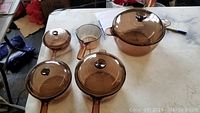 Full set of Corning Vision glass cookware with lids on a white table, includes large pot, saucepans, fry pans with brown glass lids and knobs.