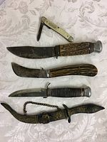 Five knives laid flat on white patterned cloth; from top: folding pocket knife with faux mother-of-pearl handle, curved utility knife with stag handle, large hunting knife with stag handle and metal pommel, fixed-blade knife with stacked leather handle and metal pommel, decorative dagger in ornate scabbard with chain.