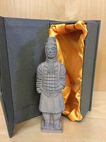 Front view of the Asian-style clay warrior figure placed inside an open gray display box with orange satin lining.