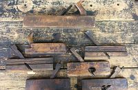 Seven antique wood planes displayed on an old wooden table, showing various sizes and wear.