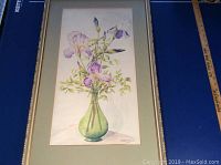 Frontal photo of framed watercolour painting showing purple irises in a green vase on a white surface.