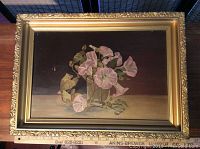 Framed oil painting on board showing overall composition of pink flowers in a clear glass vase with a dark background and beige surface, set within a carved gold frame with decorative edges.