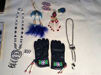 Photo showing overall collection of Indigenous beadwork items including gloves, medallion necklace, earrings, keychains, and bolo tie, arranged on a white surface.