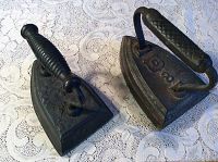 Two antique sad irons displayed on a lace tablecloth showing handles and bases.