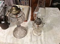 Two antique coal oil lamps on a table, one smaller finger lamp with chimney and one taller peanut-shaped lamp, both showing normal age wear.