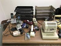 Photo of desk surface with VTech phones, Paymaster check writer, multi-tier file trays, calculator, puncher, tape dispenser, and various small office items.