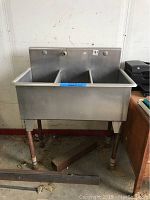 Front view of three-compartment stainless steel commercial sink with metal pipe legs against a wall.
