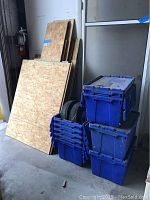 Several particle board sheets of various sizes leaning against a wall with blue plastic storage tubs stacked beside them.
