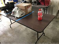 First photo of one folding table with dark wood-tone top, metal folding legs, and items placed on top including a red and clear drink bottle, cardboard boxes and other miscellaneous items.