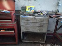 Gray metal tool chest with 3 drawers, one drawer missing, and an open bottom shelf, placed next to a red metal tool chest.
