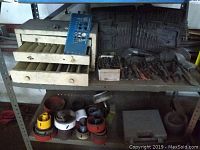 Wide view of metal shelving with metal drawers containing drill bits and hole saw attachments on lower shelf, plumbing and hand tools on top shelf.