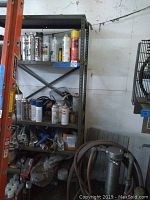 Full view of metal shelf with various spray paints, chemical bottles, tools, and miscellaneous items on three shelves.