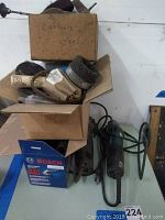 Two cardboard boxes stacked, filled with assorted grinding and sanding accessories including steel wire brushes and discs, with visible labels such as carbon steel and Bosch abrasives.