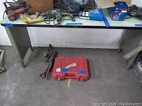 Full view of the heavy industrial table, Milwaukee branded red drill case, and metal hoist chain on the floor beneath the table.