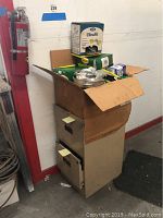 File cabinet in corner with cardboard box on top containing various items