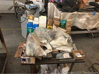 Wide view of table with steel hardware items in plastic bags and boxes on a workbench with other equipment in background.