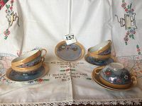 Photo showing five painted luminescent teacups and saucers arranged on an embroidered cloth with text and floral borders.