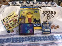 Photo of Hanukkah menorah, box of Chanukah candles, six wooden dreidels, 'I Have a Little Dreidel' story and song book, Jewish holidays book, thank you notes, all arranged on a white cloth with blue floral pattern.